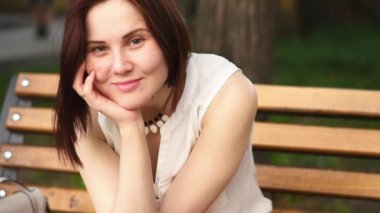 Güzel genç kadın gizemli bir şekilde, bir yaz parkta bir bankta oturan gülümsüyor. Açık havada bir kadın portresi.