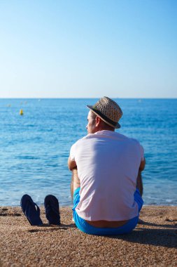 Genç bir adam sahilde oturan ve denize bakarak hasır şapkalı