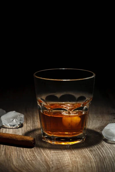 A glass of alcoholic beverage with ice cubes and a cigar on a wooden table. Whiskey in glass on black background