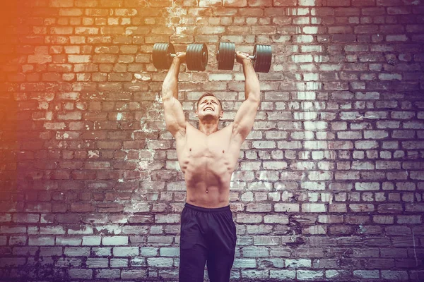 Мускулистый парень делает упражнения с гантели против кирпичной стены — стоковое фото