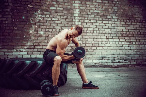 Muscular cara fazendo exercícios com halteres contra uma parede de tijolo — Fotografia de Stock