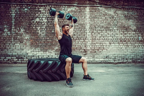 Muscular cara fazendo exercícios com halteres contra uma parede de tijolo — Fotografia de Stock