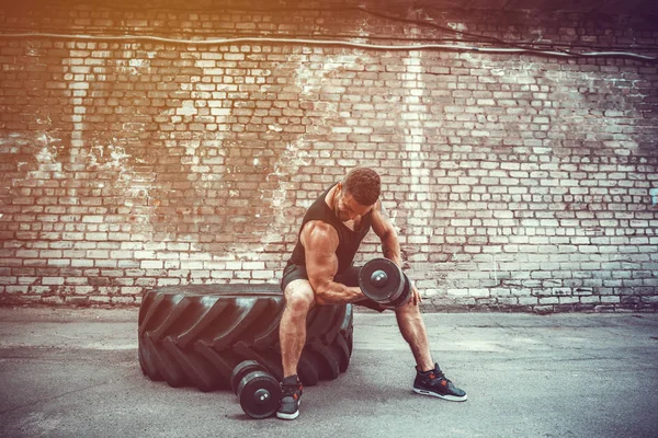 Muscular cara fazendo exercícios com halteres contra uma parede de tijolo — Fotografia de Stock