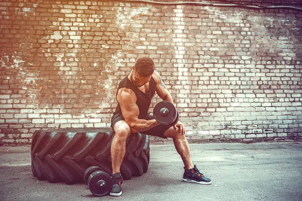 Muscular cara fazendo exercícios com halteres contra uma parede de tijolo — Fotografia de Stock