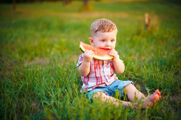 Ευτυχισμένο παιδί κάθεται στο πράσινο γρασίδι και Τρώτε καρπούζι σε εξωτερικούς χώρους στο πάρκο άνοιξη φυσικό υπόβαθρο — Φωτογραφία Αρχείου