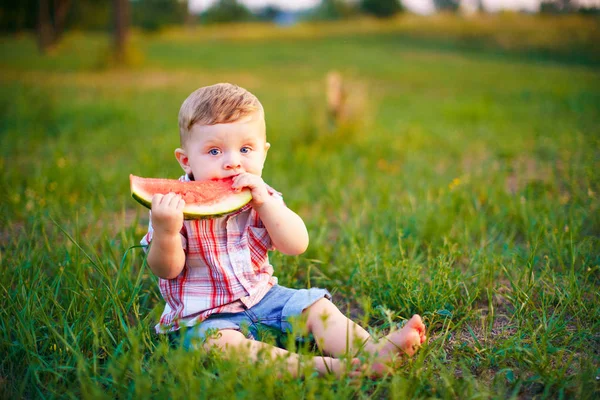 Niño feliz sentado en la hierba verde y comer sandía al aire libre en el parque de primavera contra el fondo natural —  Fotos de Stock