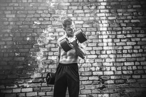 Muscular cara fazendo exercícios com halteres contra uma parede de tijolo — Fotografia de Stock