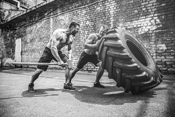 Two muscular athletes training. Muscular fitness shirtless man moving large tire — Stock Photo, Image