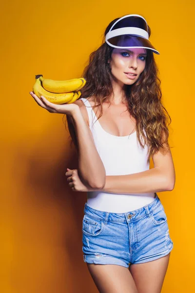 Mujer en traje de baño y pantalones cortos azules sosteniendo plátano y posando aislado sobre fondo amarillo — Foto de Stock