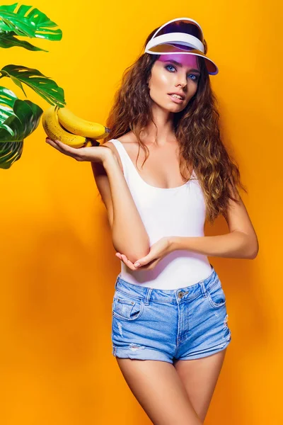 Vrouw in zwembroek en blauwe korte broek holding banaan en geïsoleerde poseren op gele achtergrond — Stockfoto