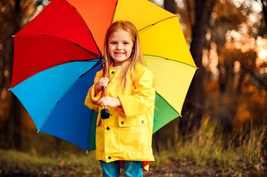 Yağmurlu ve güneşli gün bahçede oynayan renkli şemsiye ile su geçirmez ceket giyen komik şirin bebek kız