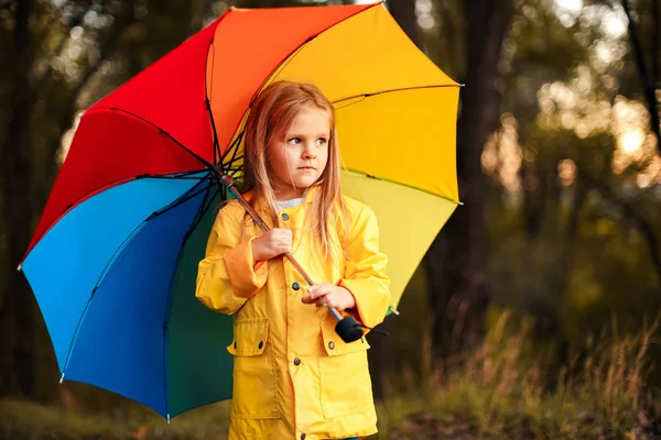 Забавная милая малышка в водонепроницаемом пальто с красочным зонтиком, играющая в саду в дождливый и солнечный день — стоковое фото