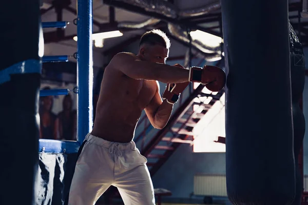 Férfi bokszoló edzés boxzsákkal a sötét sportcsarnokban. — Stock Fotó