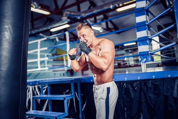 Male boxer training with punching bag in dark sports hall.