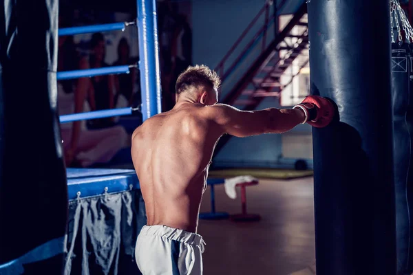 Male boxer training with punching bag in dark sports hall.