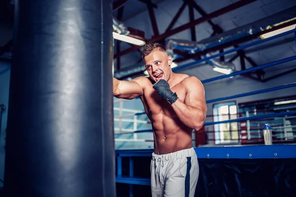 Mužský boxer trénink s boxovací taškou v tmavé sportovní hale. — Stock fotografie