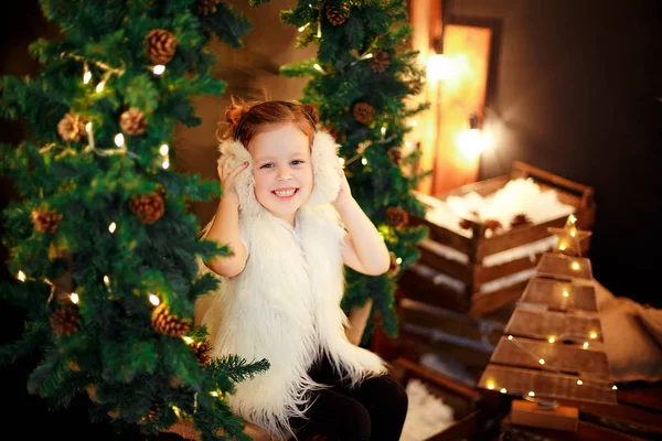 Cute little girl in fur earmuffs sitting near Christmas tree — Stock Photo, Image