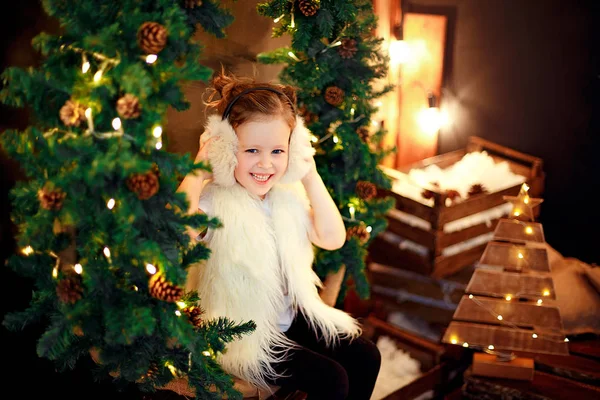 Söt liten flicka i päls öronmuffar sitter nära julgran — Stockfoto