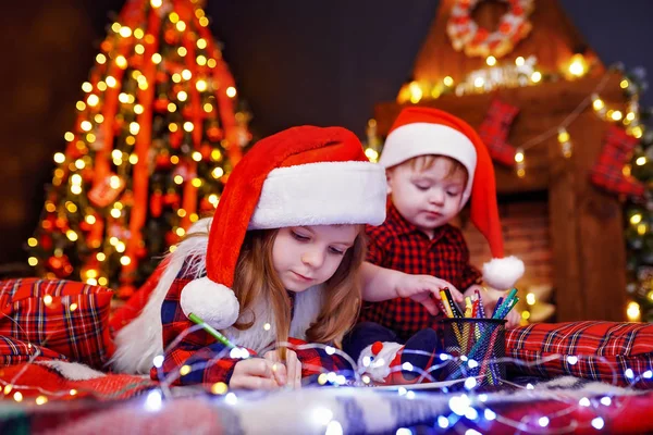 Menina engraçada em chapéu de Papai Noel escreve carta para Papai Noel e seu irmão liitle — Fotografia de Stock