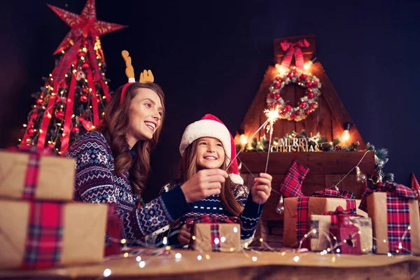 Helgdagar, familj och människor koncept. Lycklig mamma och liten flicka i santa helper hat med tomtebloss i händer, presentaskar på förgrunden ha roligt — Stockfoto