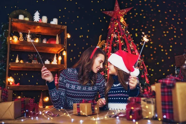 Helgdagar, familj och människor koncept. Lycklig mamma och liten flicka i santa helper hat med tomtebloss i händer, presentaskar på förgrunden ha roligt — Stockfoto