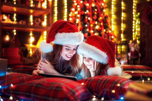 Uśmiechnięta Rodzina Matka i córka w kapeluszach Mikołaje i piżamy oglądania zabawne wideo lub wybierając prezenty na cyfrowy tablicowy — Zdjęcie stockowe