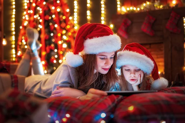 Leende familjen mor och dotter i santas hattar och pyjamas titta rolig video eller välja presenter på digitala tablett — Stockfoto