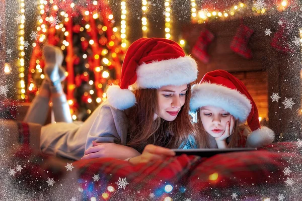 Leende familjen mor och dotter i santas hattar och pyjamas titta rolig video eller välja presenter på digitala tablett — Stockfoto