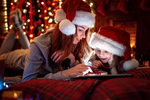 Leende familjen mor och dotter i santas hattar och pyjamas titta rolig video eller välja presenter på digitala tablett — Stockfoto