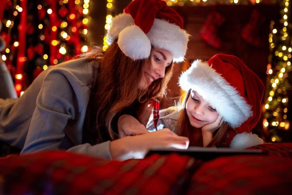 Leende familjen mor och dotter i santas hattar och pyjamas titta rolig video eller välja presenter på digitala tablett — Stockfoto