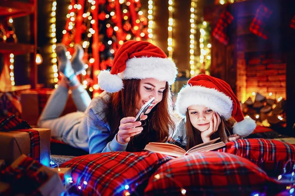 Jonge moeder met haar dochtertje lezen van een boek tijdens de vergadering onder gedecoreerde kerstboom op de verdieping — Stockfoto