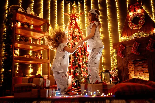 Glada små flickor bär jul pyjamas spelar en öppen spis i ett mysigt mörka vardagsrum på julafton. — Stockfoto