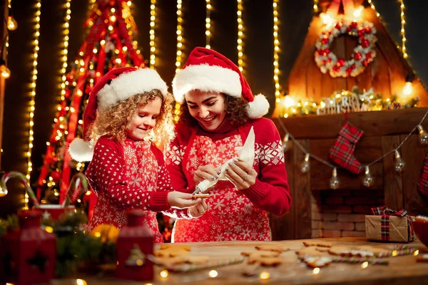 God jul och god jul. Glada söta lockigt lilla flicka och hennes äldre syster i santas hattar matlagning julkakor. — Stockfoto