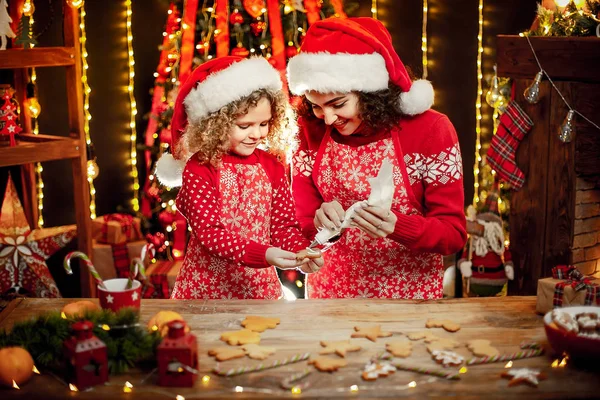 Wesołych Świąt i Wesołych Świąt. Wesoły ładny kręcone mała dziewczynka i jej starsza siostra w kapeluszach santas gotowania ciasteczka świąteczne. — Zdjęcie stockowe