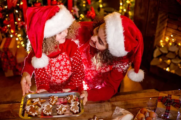 Wesołych Świąt i Wesołych Świąt. Wesoły ładny kręcone mała dziewczynka i jej starsza siostra w kapeluszach santas gotowania ciasteczka świąteczne. — Zdjęcie stockowe