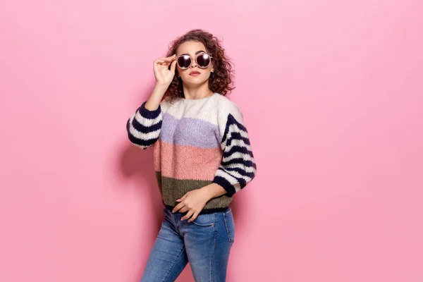 Menina bonita com cabelo encaracolado posando em camisola elegante, jeans e óculos de sol no fundo pastel rosa . — Fotografia de Stock