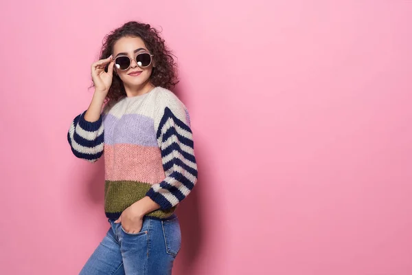 Menina bonita feliz com sorriso incrível posando em camisola elegante, jeans e óculos de sol no fundo pastel rosa . — Fotografia de Stock