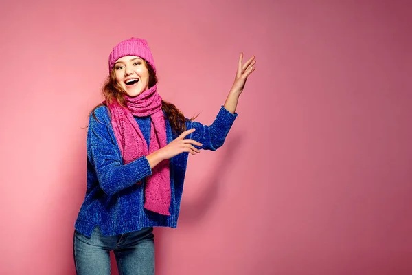 Jovem mulher moderna vestindo suéter azul e chapéu rosa, e cachecol posando, fazendo expressão facial engraçada . — Fotografia de Stock