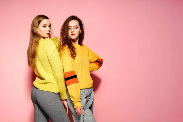 Två vackra sexiga leende underbara tjejer tittar på kameran. Heta kvinnor står i eleganta gula tröjor, på rosa bakgrund. — Stockfoto