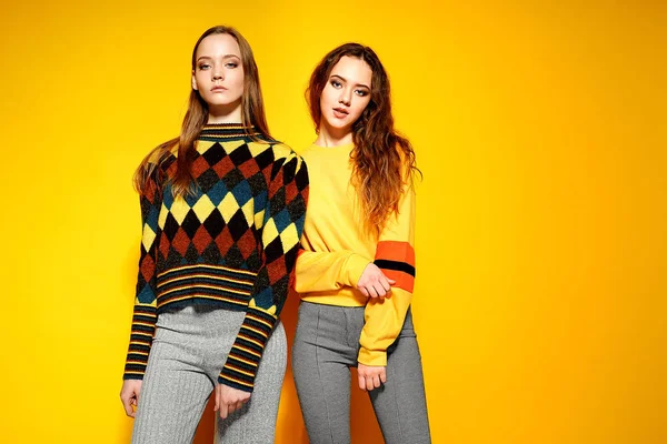 Två vackra sexiga leende underbara tjejer tittar på kameran och poserar i snygga tröjor på gul bakgrund. — Stockfoto