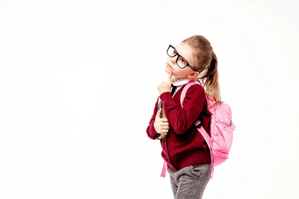 Niño con mochila. Chica en camisa blanca y pantalones grises, gafas redondeadas, sostener un libro, bolso de la escuela y posar como modelo señala el dedo hacia arriba. Aislado sobre blanco — Foto de Stock