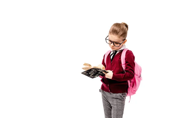 Παιδί με τσάντας. Κορίτσι με λευκό πουκάμισο και γκρι παντελόνι, στρογγυλεμένες γυαλιά, κρατήστε ένα βιβλίο, σχολική τσάντα και ποζάρει σαν μοντέλο δείχνει δάχτυλο επάνω. Απομονωμένα σε λευκό — Φωτογραφία Αρχείου