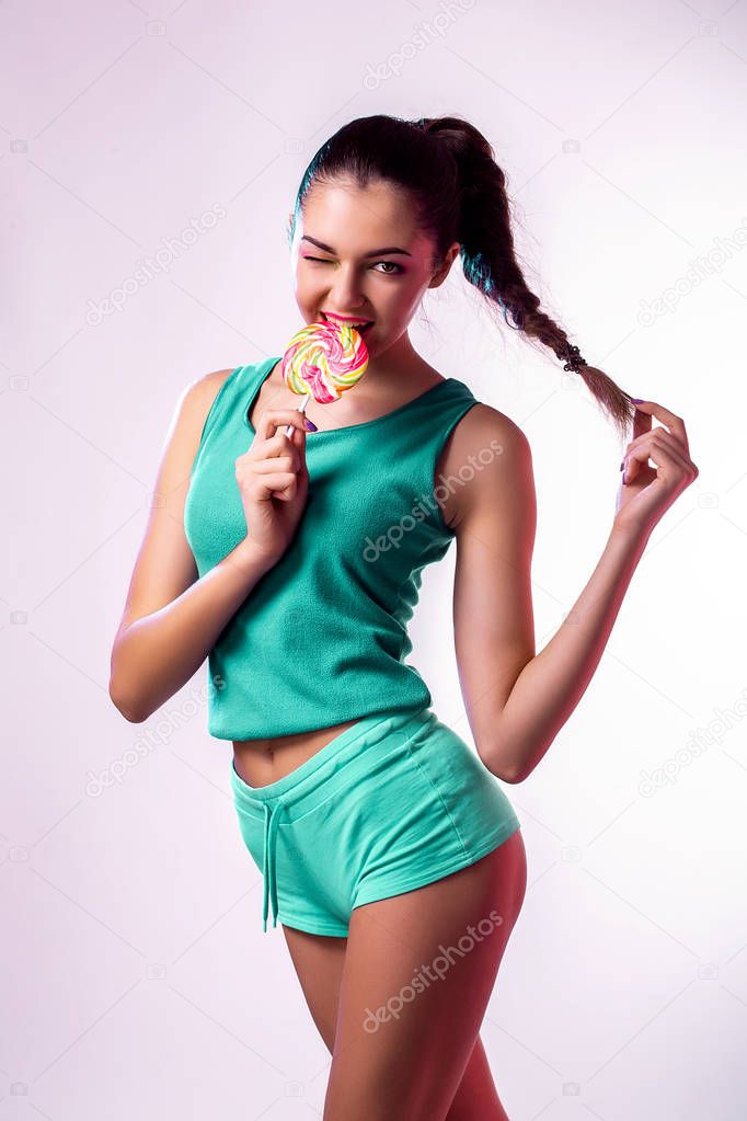 Funny young girl in green jumpsuit holding lollipop and posing for camera onwhite background.