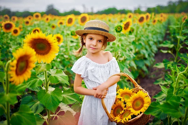 Menina em um vestido branco, um chapéu de palha com uma cesta cheia de girassóis sorrindo para a câmera em um campo de girassóis — Fotografia de Stock