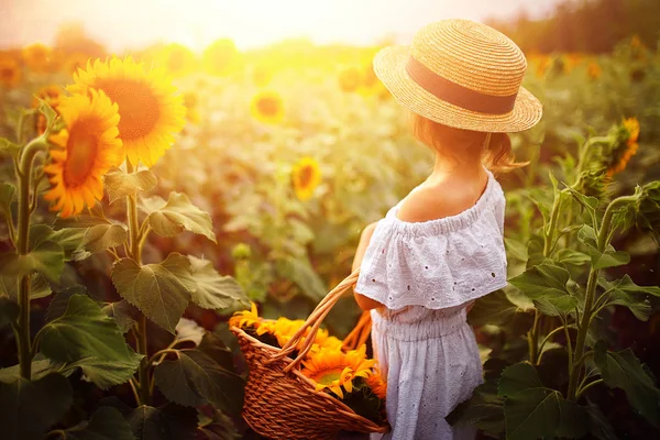 Petite fille dans une robe blanche, un chapeau de paille avec un panier plein de tournesols souriant à la caméra dans un champ de tournesols — Photo
