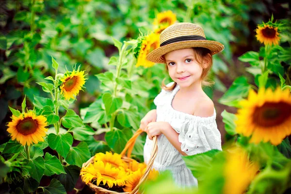 Kleines Mädchen in weißem Kleid, Strohhut mit einem Korb voller Sonnenblumen, die in einem Sonnenblumenfeld in die Kamera lächeln — Stockfoto