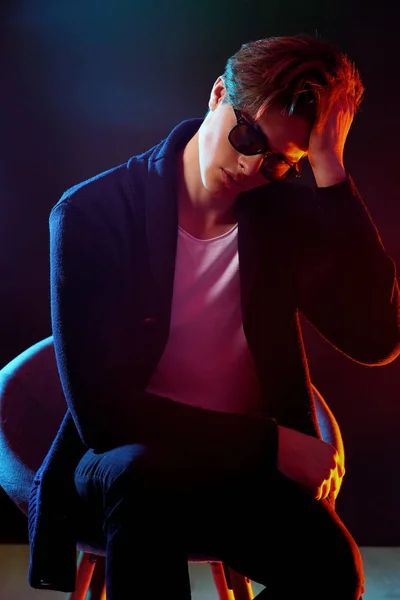 Uomo fresco ed elegante in giacca nera e occhiali da sole. Modello maschile di alta moda in luminose luci al neon colorate in posa su sfondo nero. Concetto di art design — Foto Stock