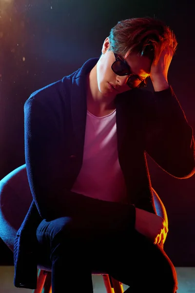 Cool stylish man in black jacket and sunglasses. High Fashion male model in colorful bright neon lights posing on black background. Art design concept