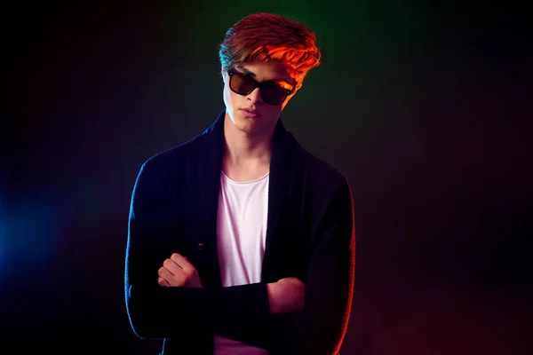 Cool stylish man in black jacket and sunglasses. High Fashion male model in colorful bright neon lights posing on black background. Art design concept