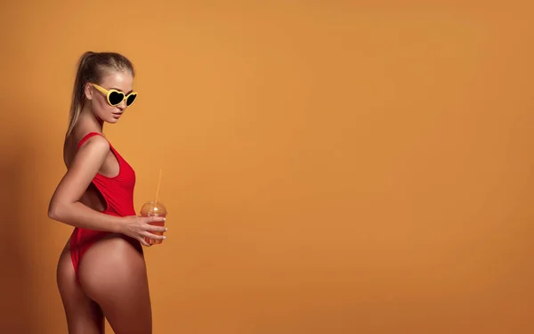 Lindo jovem em roupa de banho e óculos de sol segurando vidro de bebida fresca, enquanto em pé sobre amarelo, laranja fundo quente — Fotografia de Stock